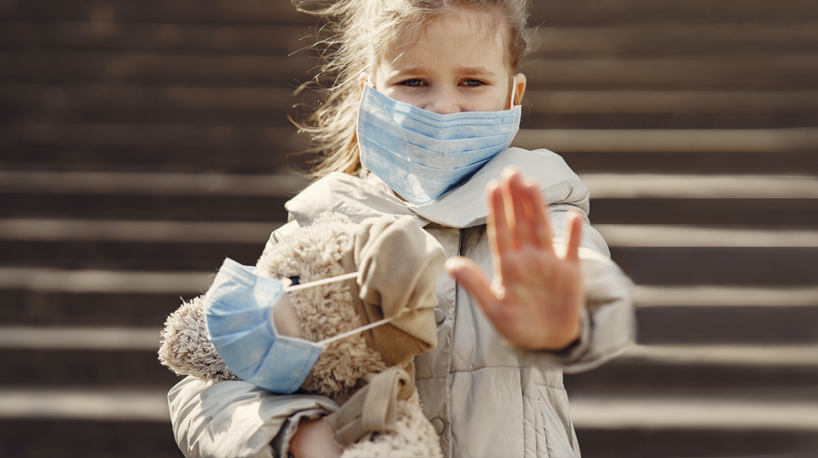 Dziewczynka z misiem, którego przytula lewą ręką. Dziewczynka i miś mają maseczki. Lewa ręka dziewczynki jest wyciągnięta w przód, otwartą dłonią.