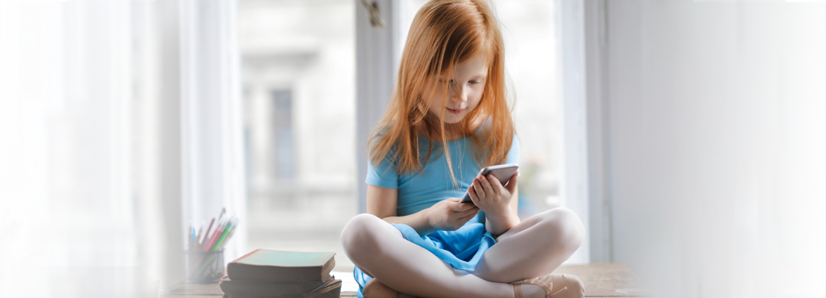 Dziewczynka siedząca ze skrzyżowanymi nogami na tle okna, przeglądająca telefon komórkowy.