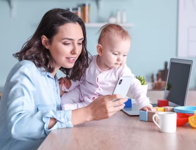 Mama z niemowlakiem, trzyma komórkę, przed nią komputer